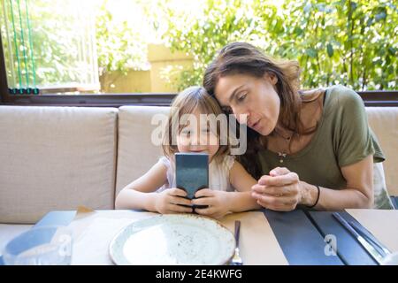 four years age blonde girl with mobile in her hands, smiling and watching with woman mother sitting in restaurant Stock Photo