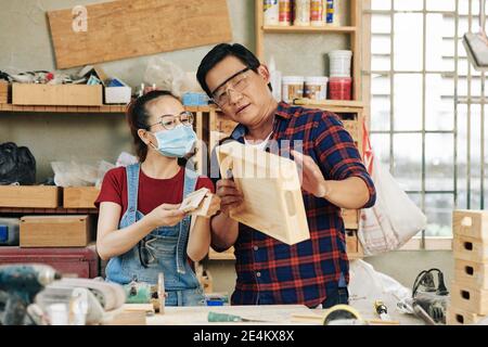 Experienced carpenter in goggles discussing quality of wooden drawer made by his apprentice Stock Photo