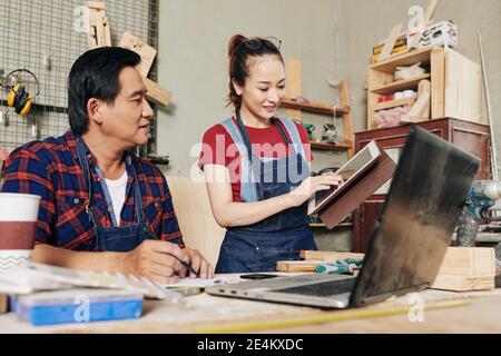 Smiling young female carpenter showing coworker tablet computer with furniture item draft on ther screen that she was working on Stock Photo