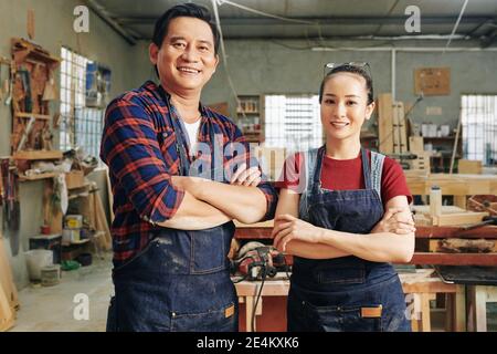 Cheerful mature carpenters and his apprentice standing in workshop in denim aprons, folding arms and looking at camera Stock Photo