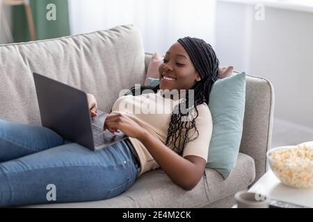 Joyful african lady laying on couch at home, using laptop Stock Photo