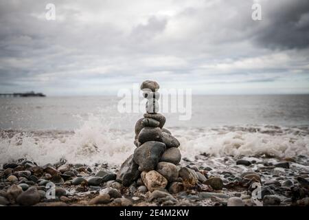 Pile of stones stacked up balancing sea shore ocean lapping up pebbles tower grey dramatic storm clouds gather above waves crashing harmony zen sea Stock Photo