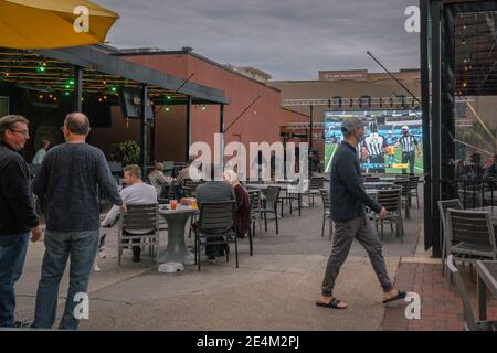 Bar with outdoor seating during Covid-19 pandemic. Patrons are socially distancing & some are masked while watching a football game. Stock Photo