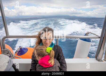 mother, smiling and looking at, embracing her daughter, four years old blonde girl with pigtail, sitting on the stern of motorboat sailing from Tarifa Stock Photo
