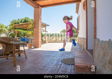 side view of four years old blonde girl with pink shirt jumping, or taking a great leap, from the exterior doorway to the porch of the house Stock Photo