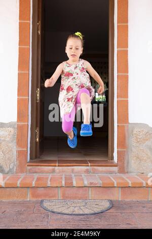 front view of four years old blonde girl with dress and pigtail jumping, or taking a great leap, from the exterior doorway of the house Stock Photo