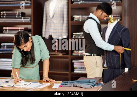 Atelier manager talking on phone with customer and writing in planner when her colleague making bespoke suit client in background Stock Photo