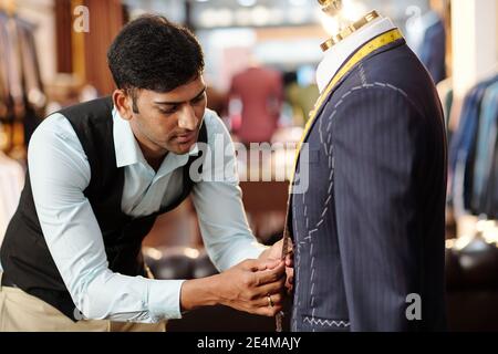 Talented young Indian tailor working on bespoke suit on mannequin, and measuring parts with tape measure Stock Photo