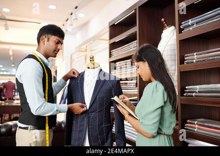 Young Indian tailor working on bespoke jacket and measuring details when his assistant taking notes in planner Stock Photo