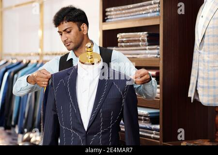 Serious talented young Indian tailor checking bespoke jacket on mannequin when working on suit for client Stock Photo