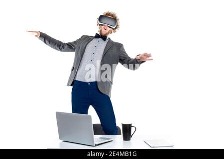Portrait of curly-haired businessman using vr glasses while standing on chair at table with gadgets and cup of coffee. Stock Photo