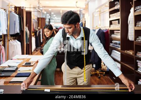Pensive handsome Indian tailor measuring fabric on table in atelier when manager checking catalogues Stock Photo