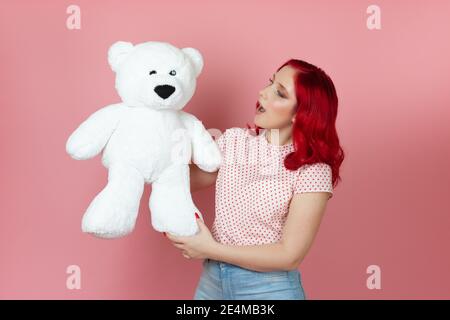 close-up a surprised, shocked young woman with an open mouth and red hair holds a large white teddy bear isolated on a pink background Stock Photo