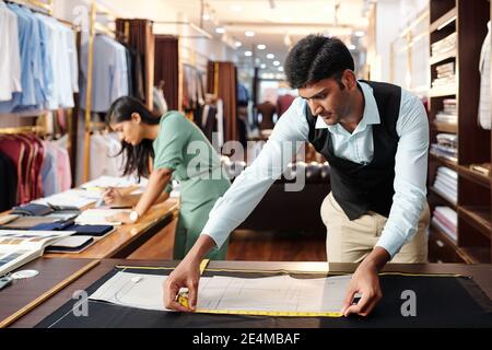 Concentrated young Indian tailor measuring sewing pattern of jacket for client Stock Photo