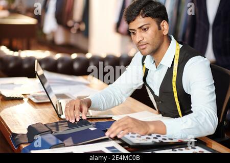 Pensive young Indian tailor checking catalog and choosing fabric for making suit Stock Photo