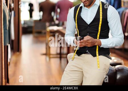 Cropped image of young tailor leaning on chair and checking text messages in smartphone during break Stock Photo