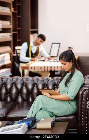 Young Indian atelier manager sitting on sofa and reading articles and e-mails from clients on tablet computer when tailor calculating fabric consumpti Stock Photo