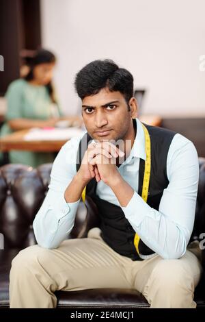 Portrait of young serious Indian tailor sitting on sofa in atelir and looking at camera Stock Photo