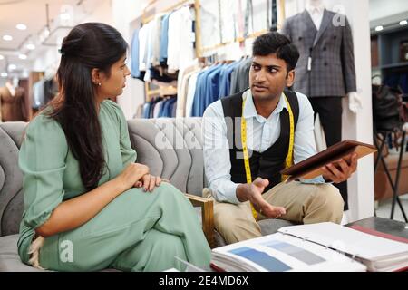 Tailor and client sitting on sofa in atelier and discussing fabric for bespoke jacket Stock Photo