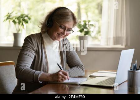 Smiling mature woman wearing headphones writing notes, using laptop Stock Photo