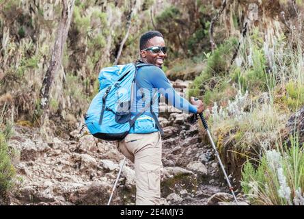 Portrait of a cheerfully smiling African-American Ethnicity young man in sunglasses. He having a walk with a backpack using trekking poles to Mweka ga Stock Photo