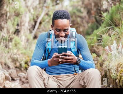 Young African ethnicity man video chatting via cellphone with somebody and cheerfully laughing as he having a hiking walk in the forest. Happy people, Stock Photo
