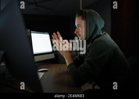 Young internet hacker in hood sitting at monitors Stock Photo