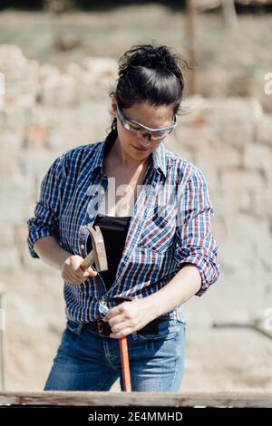 Female constructer wearing protective goggles and using hammer and spike Stock Photo