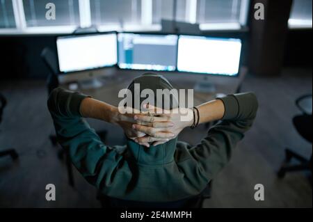 Male hacker in hood sitting at screens, back view Stock Photo
