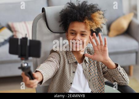 Portrait of smiling African-American boy waving at camera while holding selfie stick and video blogging from home, copy space Stock Photo