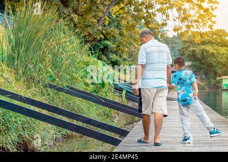 Back view of Asian grandfather and grandchild wear protective face mask to prevent Coronavirus (COVID-19) walking in a nature path on wooden bridge al Stock Photo