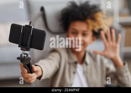 Portrait of smiling African-American boy waving at camera while holding selfie stick with smartphone and video blogging from home, focus on foreground