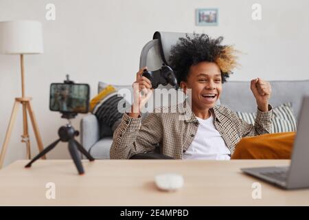 Portrait of excited African-American boy cheering while playing video games at home and online streaming, young gamer or blogger concept, copy space