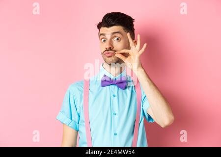 Funny young man touching his french moustache and pucker lips, looking silly at camera, standing in bow-tie and suspenders on pink background Stock Photo