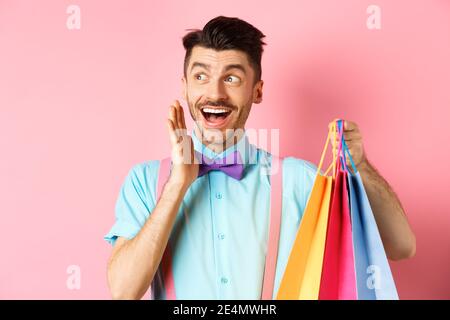 Surprised happy man screaming of joy, looking aside at logo and holding shopping bags, standing over pink background Stock Photo
