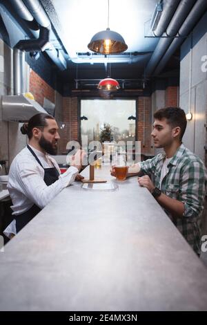 Vertical shot of a bartender talking to male customer over the counter at beer pub Stock Photo