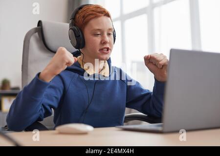 Portrait of red haired teenage boy playing video games at home and celebrating win emotionally, copy space Stock Photo