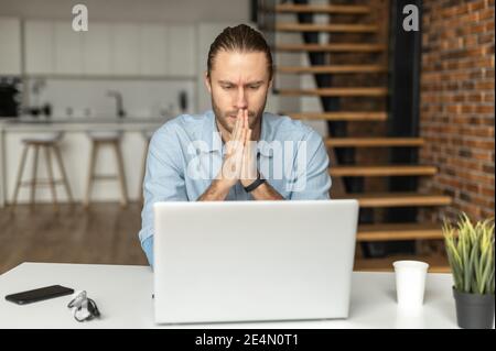 Pensive male freelancer looks at laptop screen anxiously, folded hands in prayer gest, found a mistake in a report, a worried businessman looking for Stock Photo