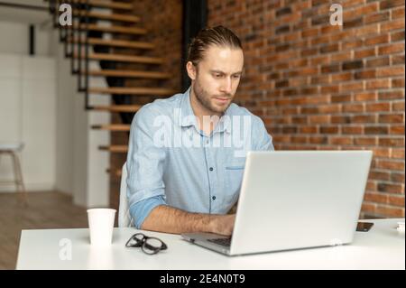 Serious intelligent male freelancer using laptop for work online sitting at the desk in the coworking space, a thoughtful guy typing on the keyboard Stock Photo