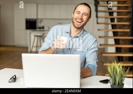 Cheerful millennial guy works on outsours, using a laptop for receiving emails, conducts correspondence with customer, develops software working Stock Photo
