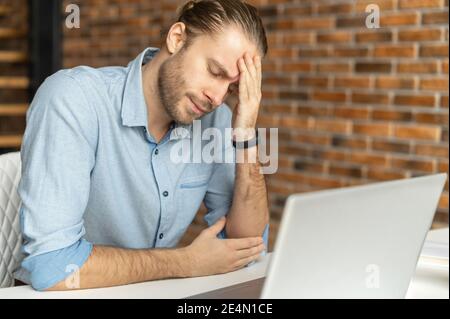 Fatigued hipster using laptop for working, a guy tired and burnout, holding head and feels headache, migraine sitting at the desk in modern office Stock Photo