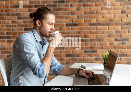 A handsome hipster drinking morning coffee, using a laptop for work and preparing to productive work day. A millennial guy works on outsours Stock Photo