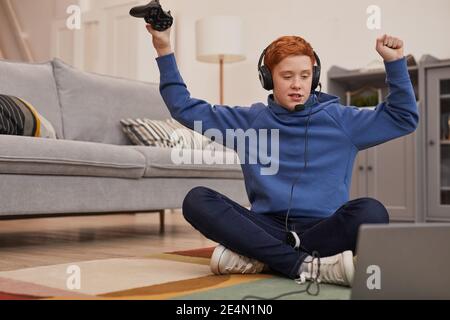 Full length portrait of excited teenage boy playing video games while sitting on floor and celebrating win, copy space