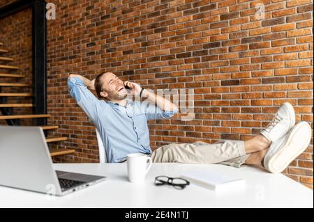 A confident hipster guy is sitting with his feet in sneakers on the table in modern office space and talking on the phone, an ambitious man resting on Stock Photo