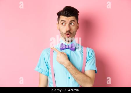Intrigued young man pointing finger left at logo, checking out promo offer, standing in bow-tie and classy outfit, pink background Stock Photo