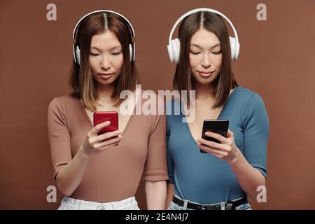 Young Asian twin sisters in casualwear and headphones listening to music and scrolling through playlists in smartphones in front of camera Stock Photo