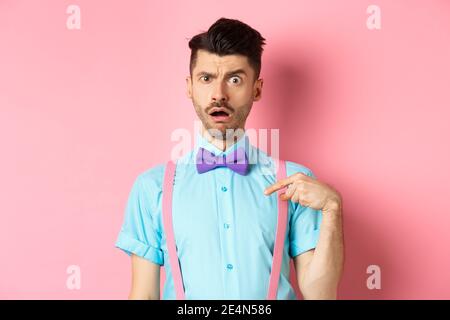 Image of confused funny guy in bow-tie and suspenders, pointing at himself as if being accused or chosen, raising eyebrow surprised, standing over Stock Photo