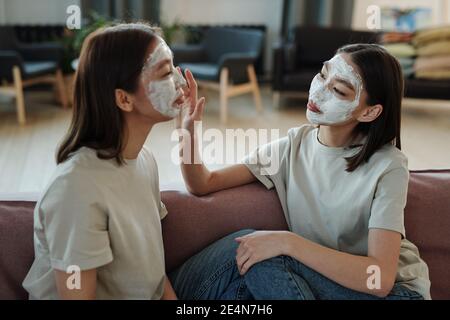 Young female twins with facial clay mask sitting on couch in front of camera at home while one of them touching face of her sister