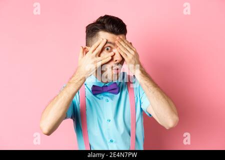 Intrigued funny guy in bow-tie, peeking through fingers at something interesting, checking out something cool, standing on pink background Stock Photo