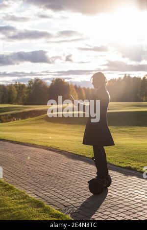 Outline of contemporary businessman in coat making selfie while standing on gyroscope against green field, trees and cloudy sky on sunny day Stock Photo